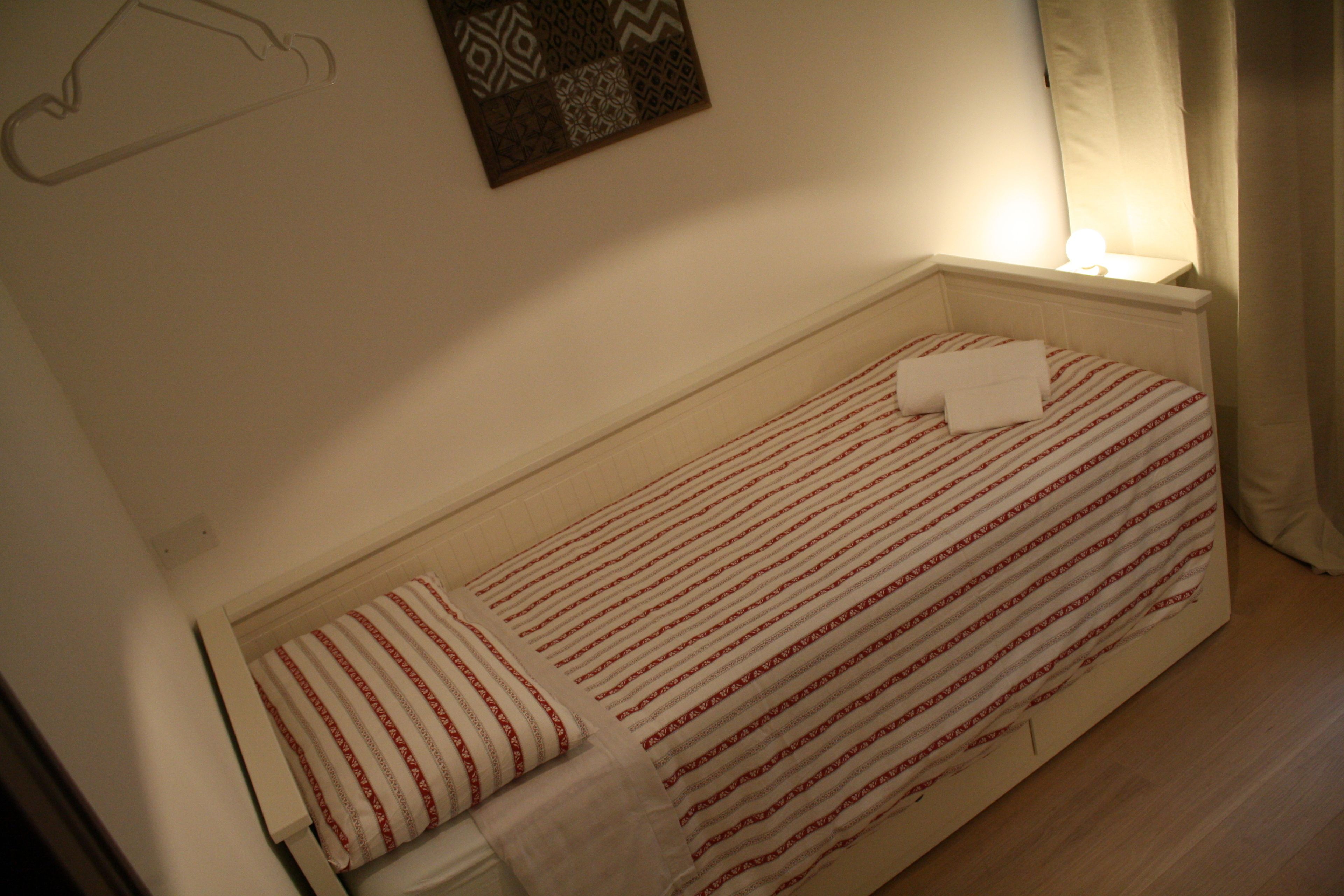 A small, single-bed room with a striped bedspread, a bedside table with a lamp, and a wall-mounted decorative item.