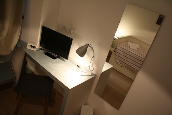 Habitación de hotel con escritorio, computadora, lámpara, cama con manta a rayas y espejo en la pared.