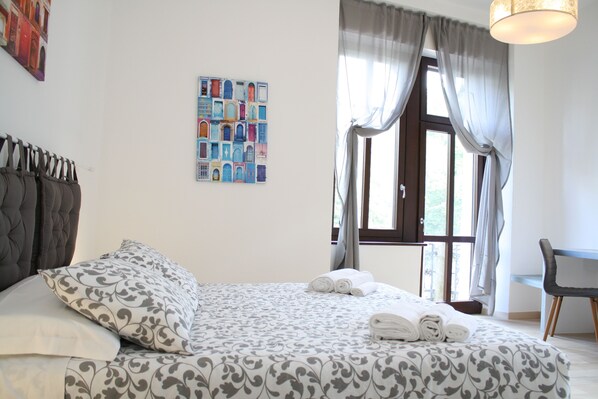 A neatly made bed with patterned bedding, a headboard, and rolled towels. A window with curtains and a view of greenery is visible.