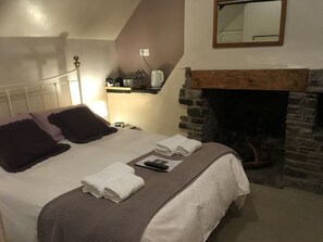 Een keurig opgemaakt bed met kussens, een nachtkastje met een lamp, een stenen openhaard en een spiegel aan de muur.