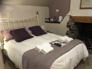 Een opgemaakt bed met witte en paarse beddengoed, een afstandsbediening en gevouwen handdoeken op een bedsprei met structuur.