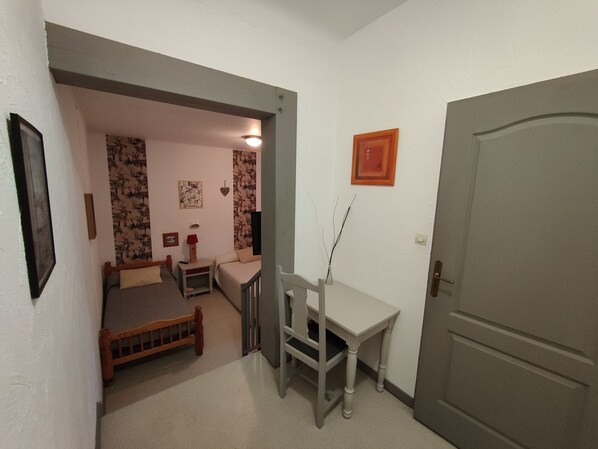 Una habitación pequeña y bien iluminada con una cama, un escritorio y una silla. Se observa una puerta a la derecha y un cuadro en la pared.