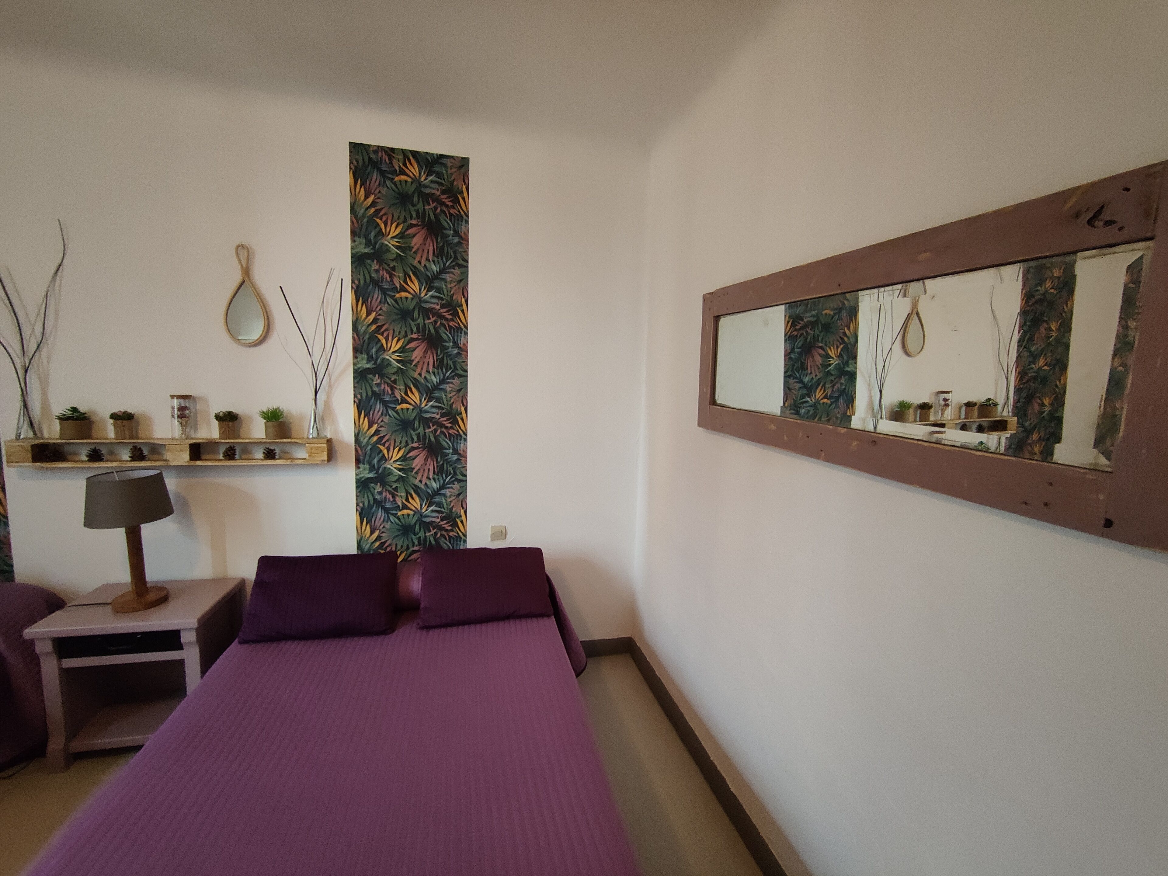 A bedroom with a bed, a nightstand, a lamp, a shelf with plants, a mirror, and a wall with a tropical leaf pattern.