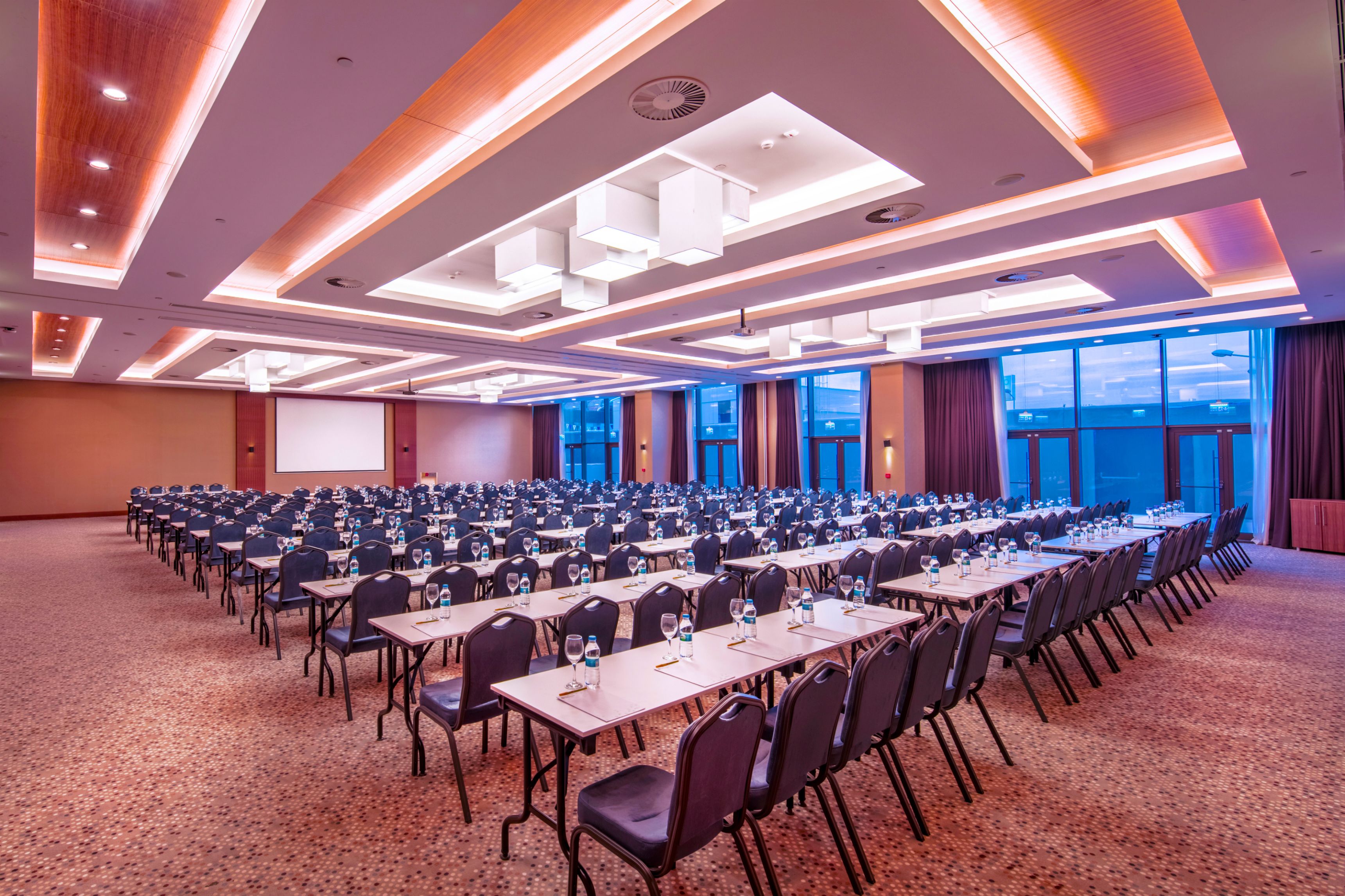 Un salón de conferencias con filas de mesas y sillas, ventanas amplias y un diseño moderno en el techo.