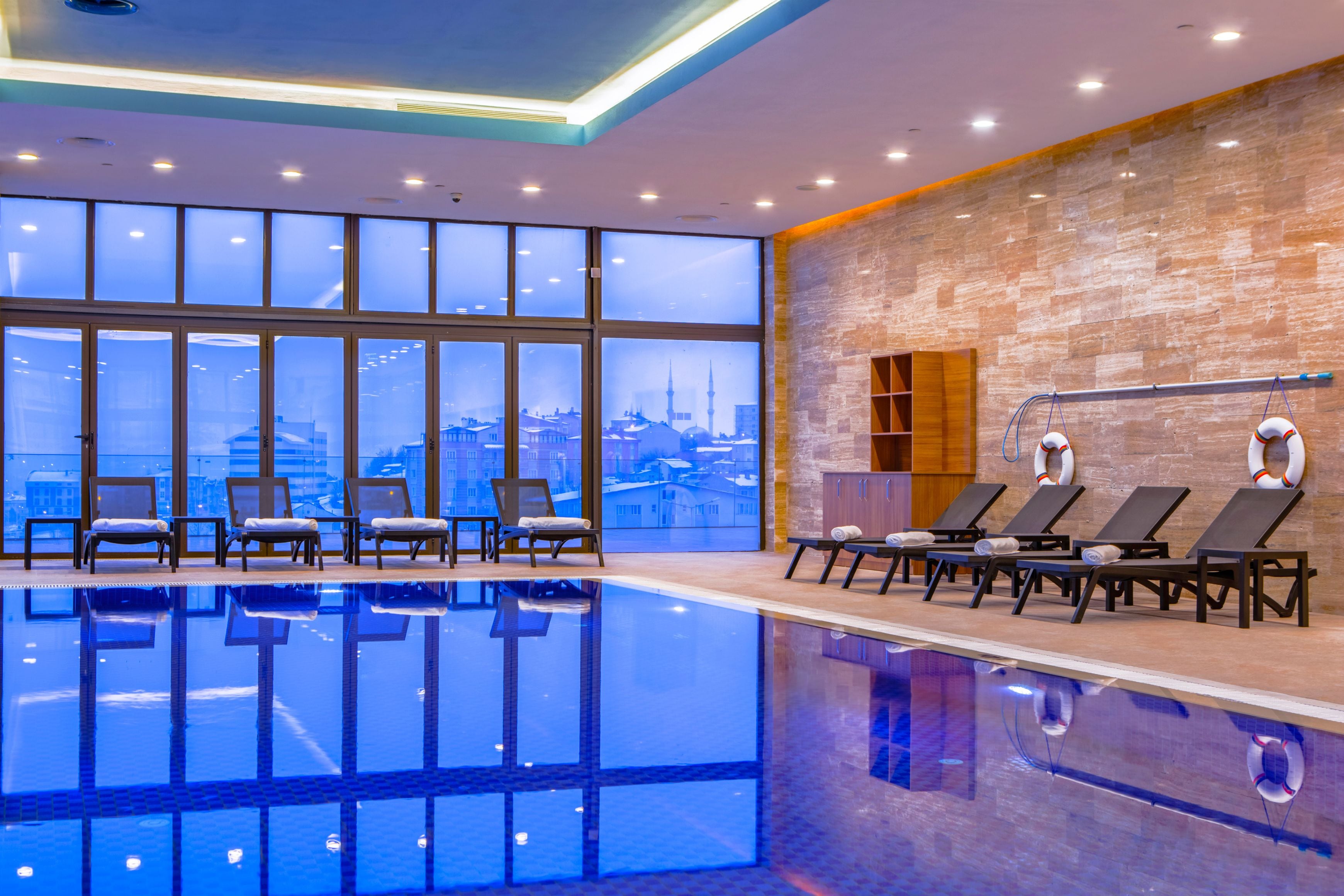 An indoor swimming pool with lounge chairs and a city view.