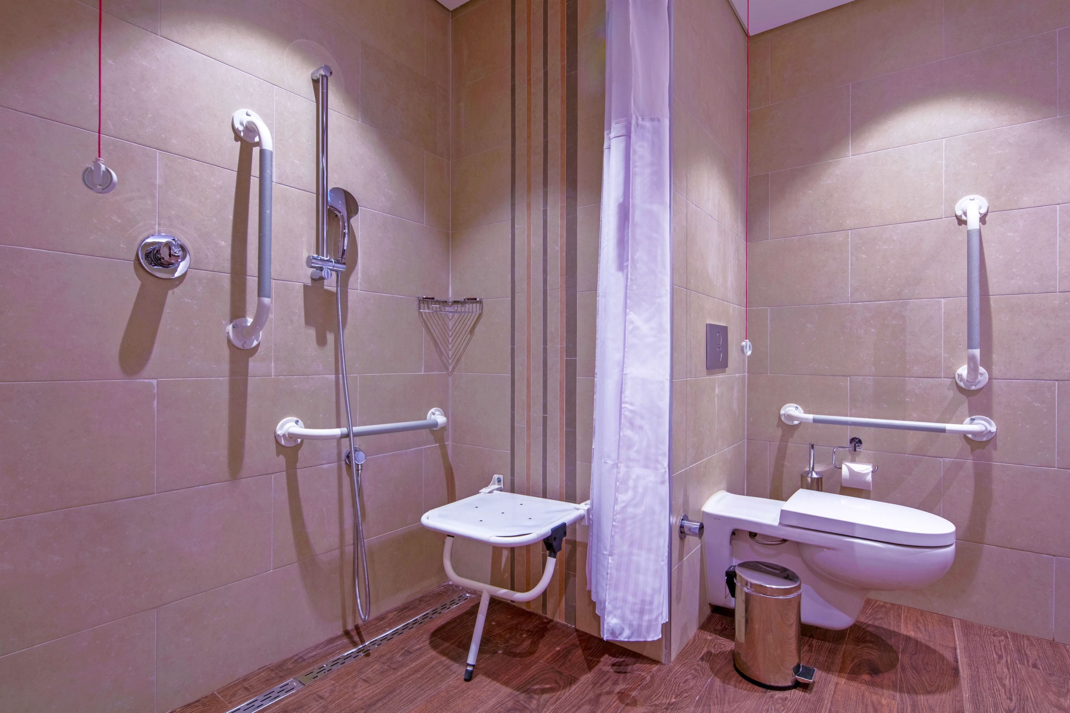 Un baño con asiento de ducha, barras de apoyo, inodoro y cubo de basura.
