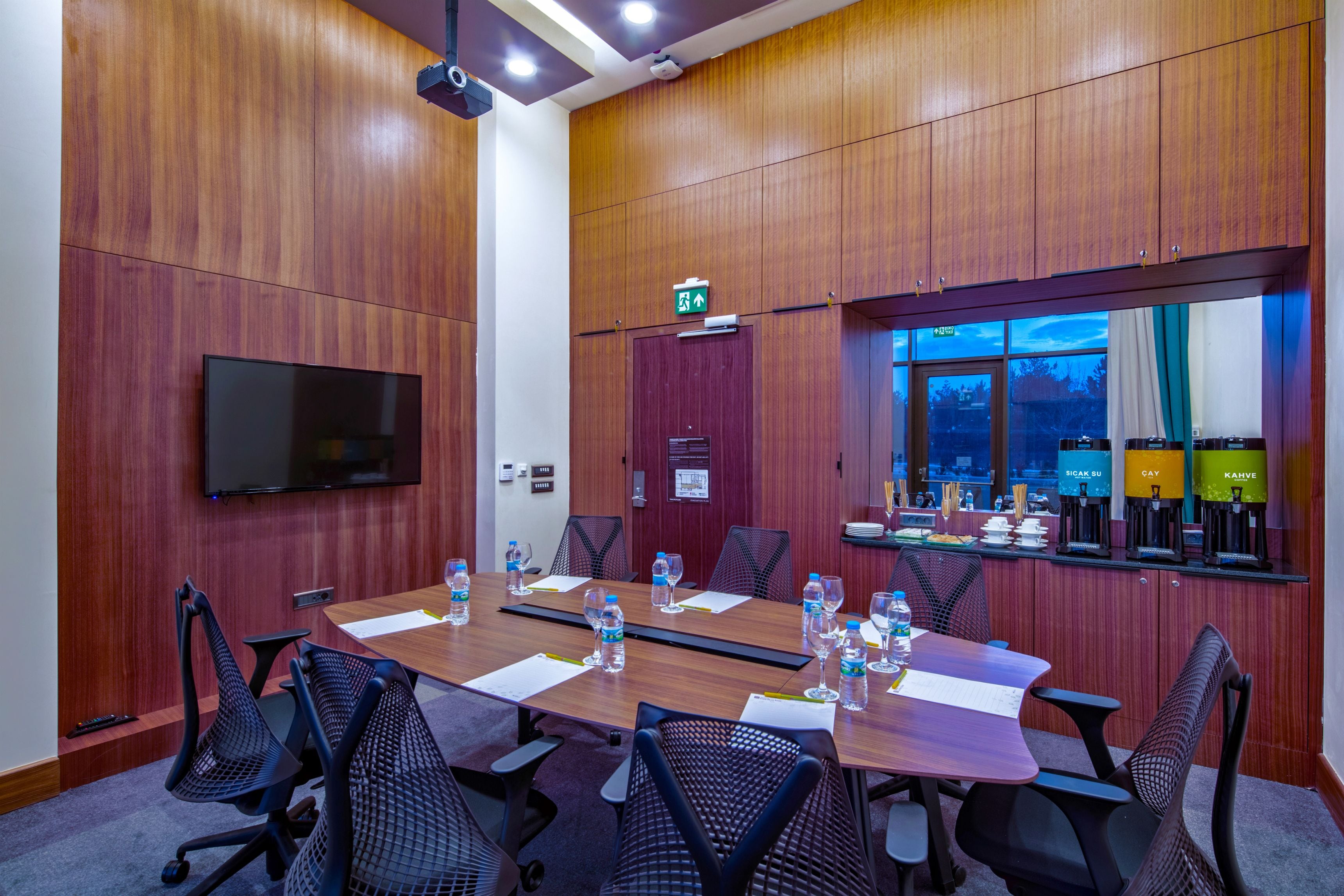 Un salón de conferencias con una larga mesa, sillas, un televisor de pantalla plana, una puerta con letrero de salida y una ventana con vistas al cielo.