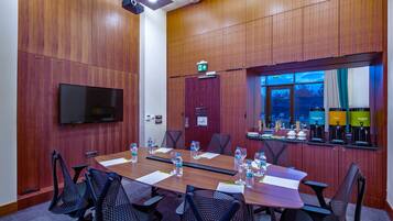 Un salón de conferencias con una larga mesa, sillas, un televisor de pantalla plana, una puerta con letrero de salida y una ventana con vistas al cielo.