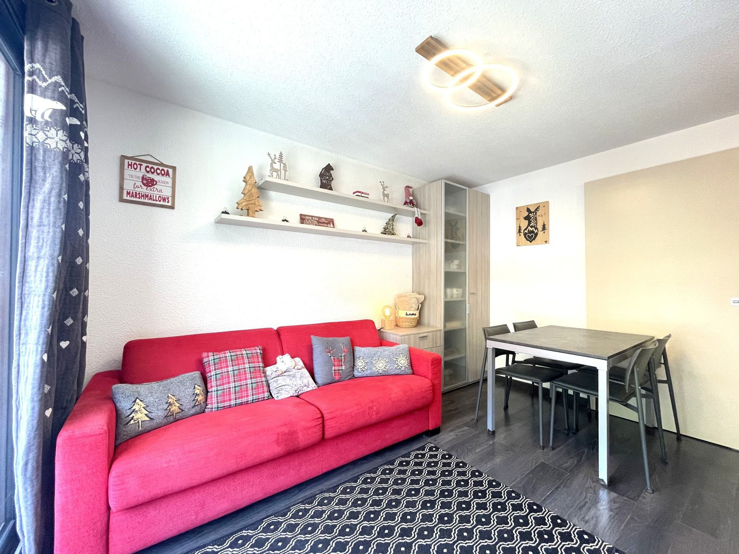 A red sofa with patterned cushions, a dining table with chairs, and shelves with various decorations.