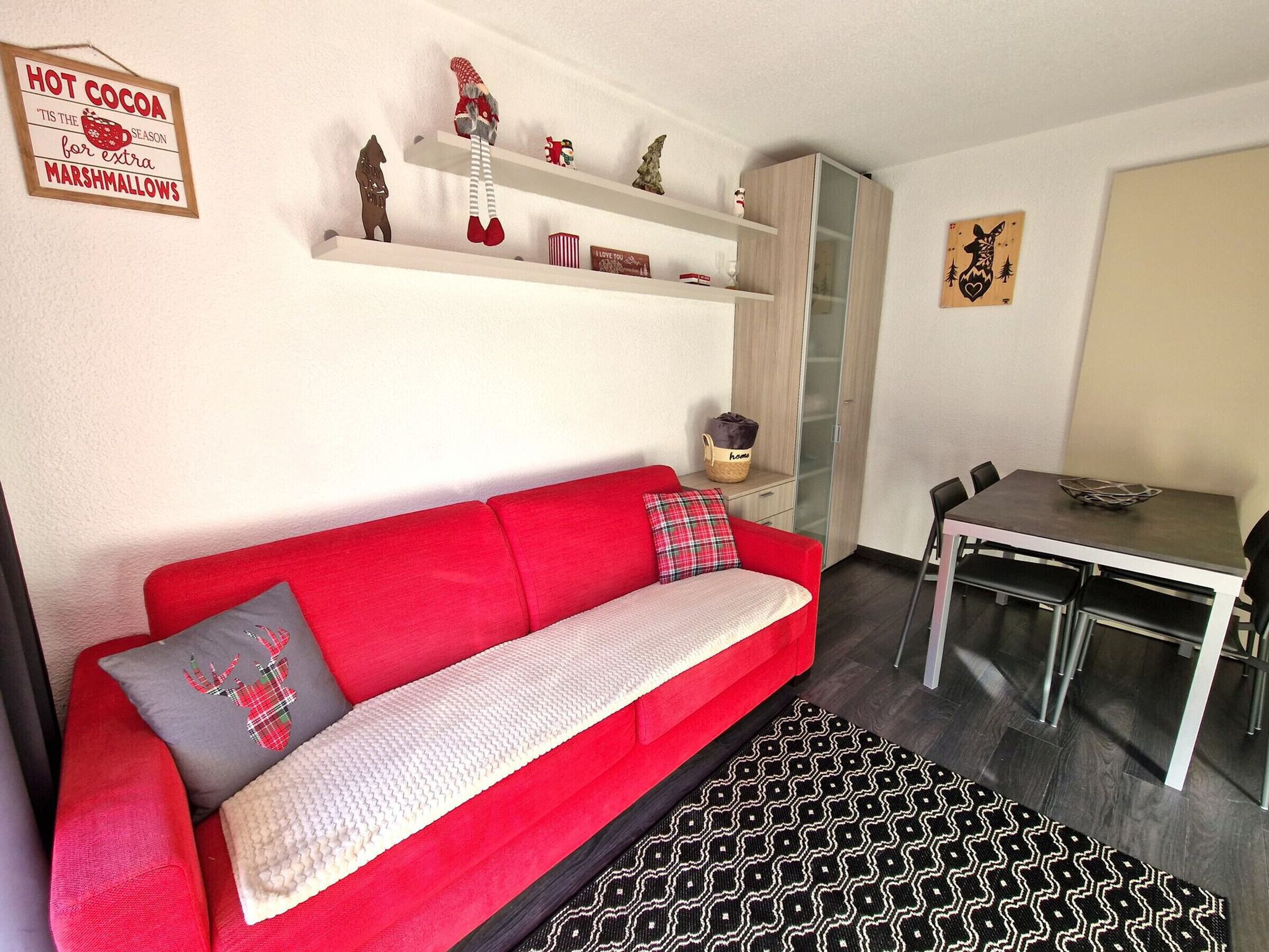 A modern living room with a red sofa, a patterned rug, a dining table with chairs, and shelves with various decorations.
