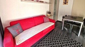 A modern living room with a red sofa, a patterned rug, a dining table with chairs, and shelves with various decorations. - Cozy 4-6 Person Apartment in Ski Resort Center with Balcony and Modern Amenities (Valfréjus)