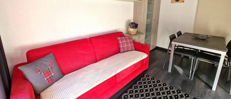 A modern living room with a red sofa, a patterned rug, a dining table with chairs, and shelves with various decorations.