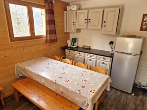 A cozy kitchen with wooden cabinets, a refrigerator, a coffee maker, and a table set with a floral tablecloth. - Spacious 4-Room Apartment for 6-8, Near Ski Slopes in Modane, France (Valfréjus)