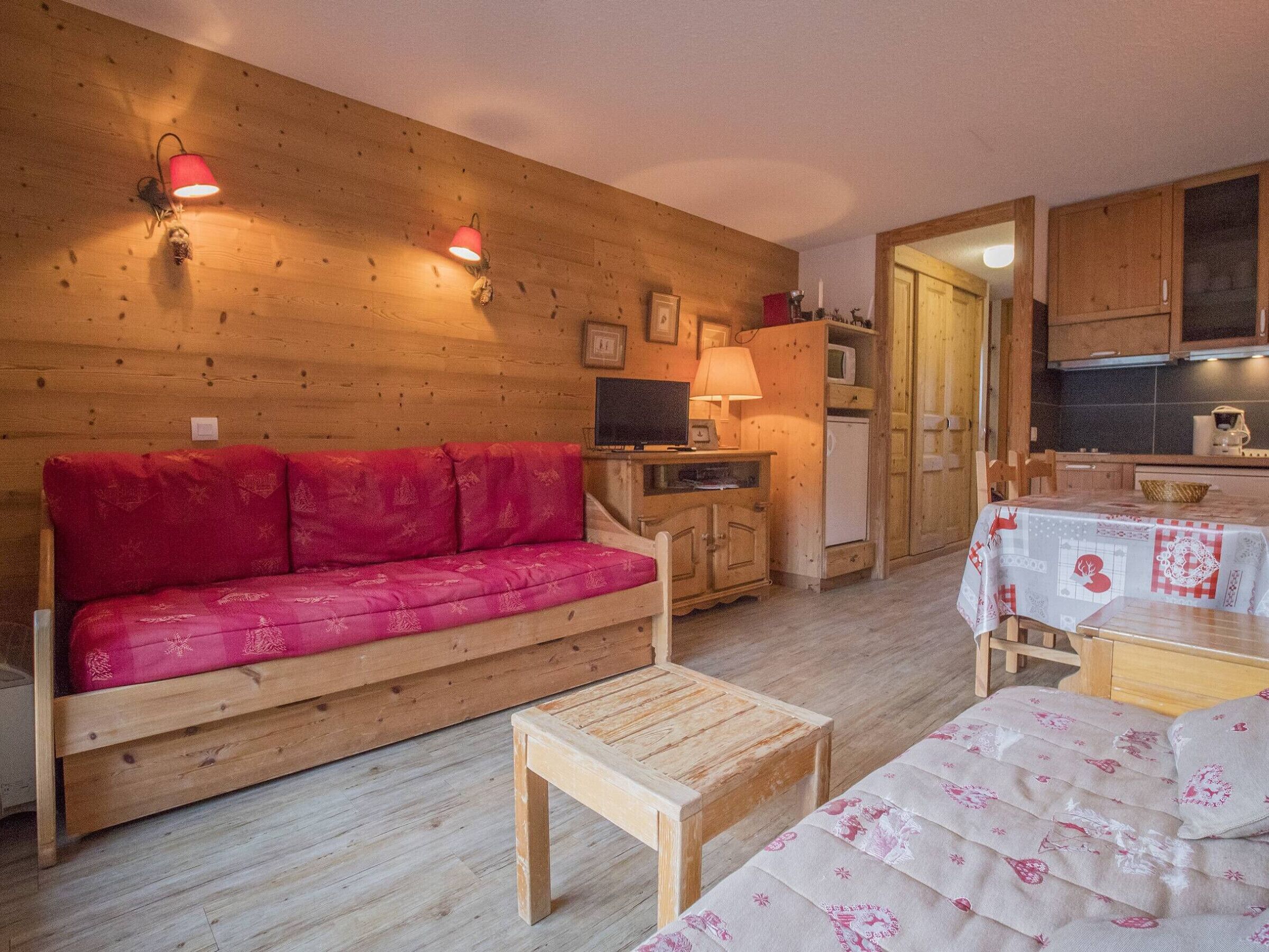 A cozy room with a wooden wall, a red sofa, a wooden table, and a kitchen area with wooden cabinets and a microwave.