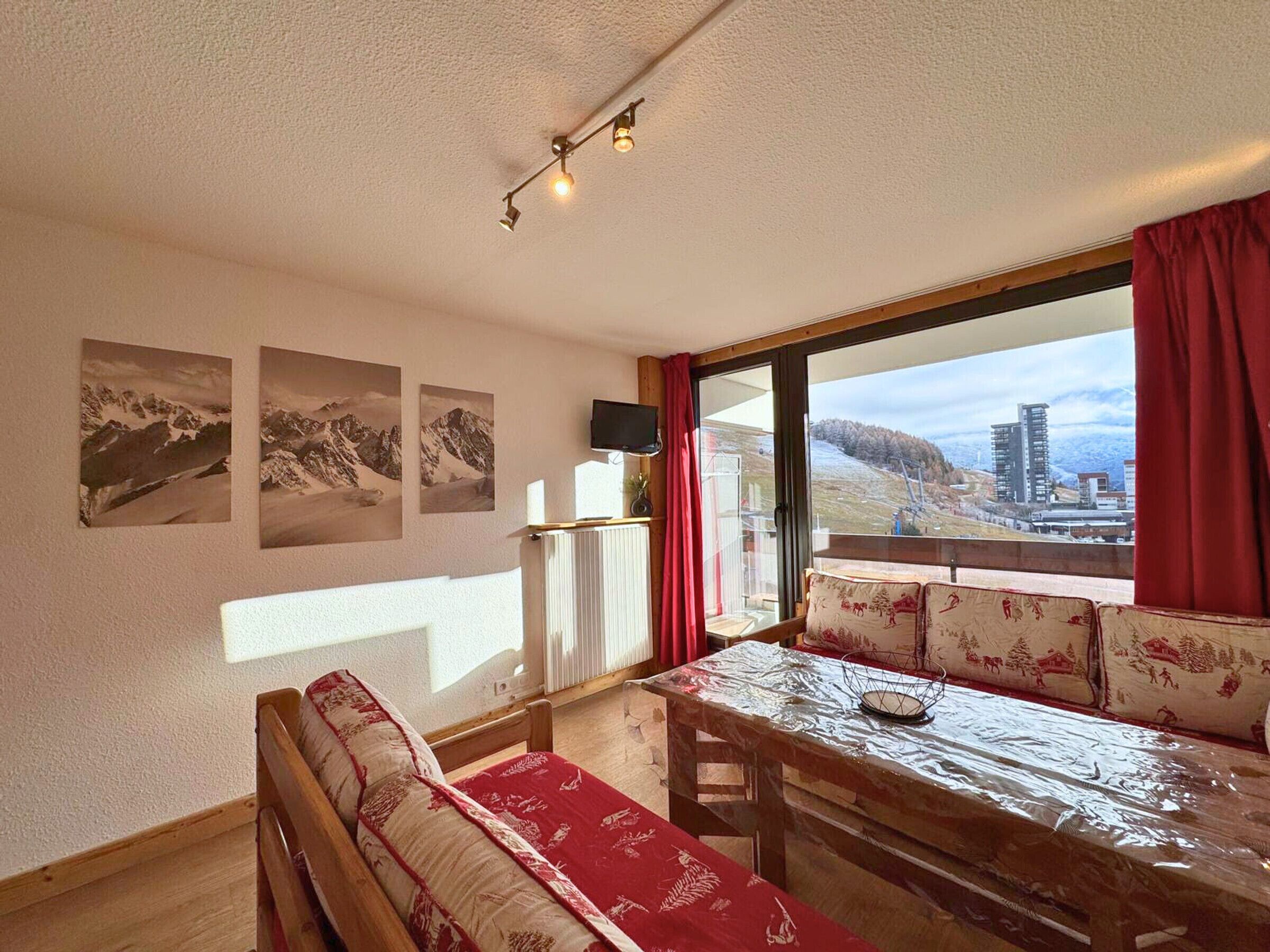 Um quarto com uma mesa de jantar, bancos, radiador e uma janela grande com vista para uma paisagem montanhosa e edifícios.