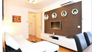 A modern living room with a sofa, a TV mounted on a wooden wall, and a small shelf with decorative items. - Apartman Trnje (Zagreb)