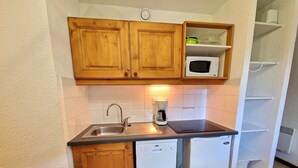 Una cocina compacta con armarios de madera, microondas integrado, cafetera, lavavajillas y una pequeña estantería.