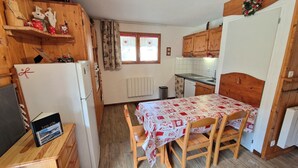 Eine kleine Küche mit Holzausstattung, einem Kühlschrank, einem Esstisch mit Stühlen und einem Fenster mit Vorhängen.