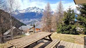 Ein Holztisch für ein Picknick in einer Berglandschaft, mit Blick auf schneebedeckte Gipfel und ein kleines Gebäude im Hintergrund.