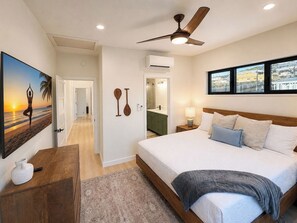 A bedroom with a bed, a ceiling fan, a painting, and a view through the window. - Brand-new Maui home with comfort and local charm! (Lahaina)