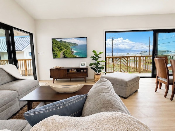 A modern living room with a sofa, a coffee table, a TV mounted on the wall, and a balcony with a view of the sea.