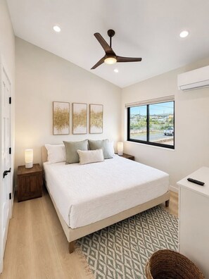 A bedroom with a bed, bedside lamps, a ceiling fan, a window with a view, and a small desk with a remote control.
