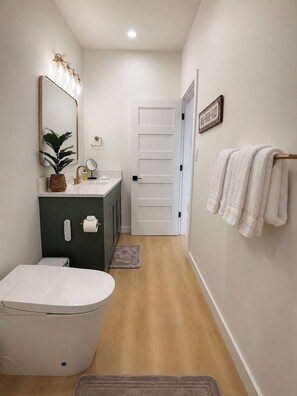 A narrow hallway with a toilet, sink, and mirror. There is a towel rack with towels and a door leading to another room. - Brand-new Maui home with comfort and local charm! (Lahaina)