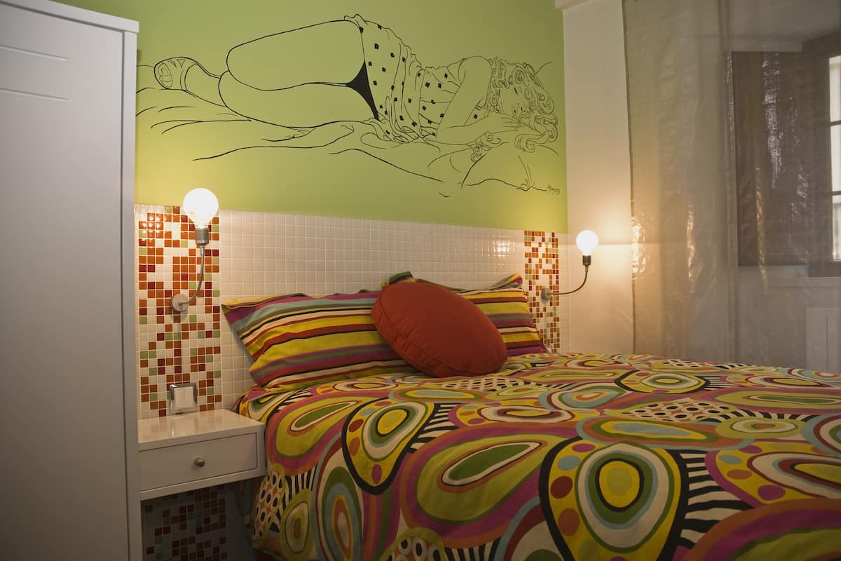 Quarto com colcha colorida, travesseiro vermelho, painel na parede e duas luminárias de cabeceira.