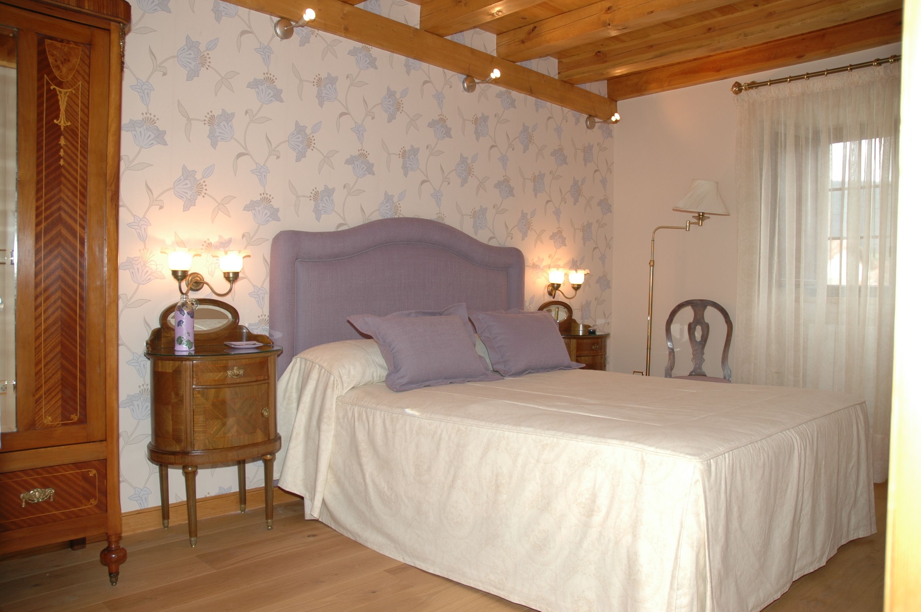 A bedroom with a large bed, wooden headboard, two bedside tables with lamps, floral wallpaper, and a wooden wardrobe.