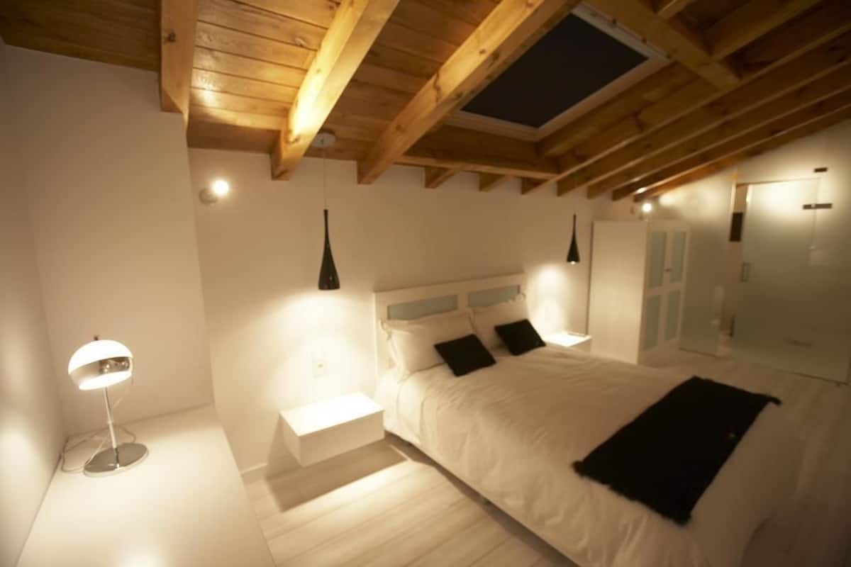 Quarto com teto de madeira, cama com roupa de cama branca, mesa de cabeceira e abajur.