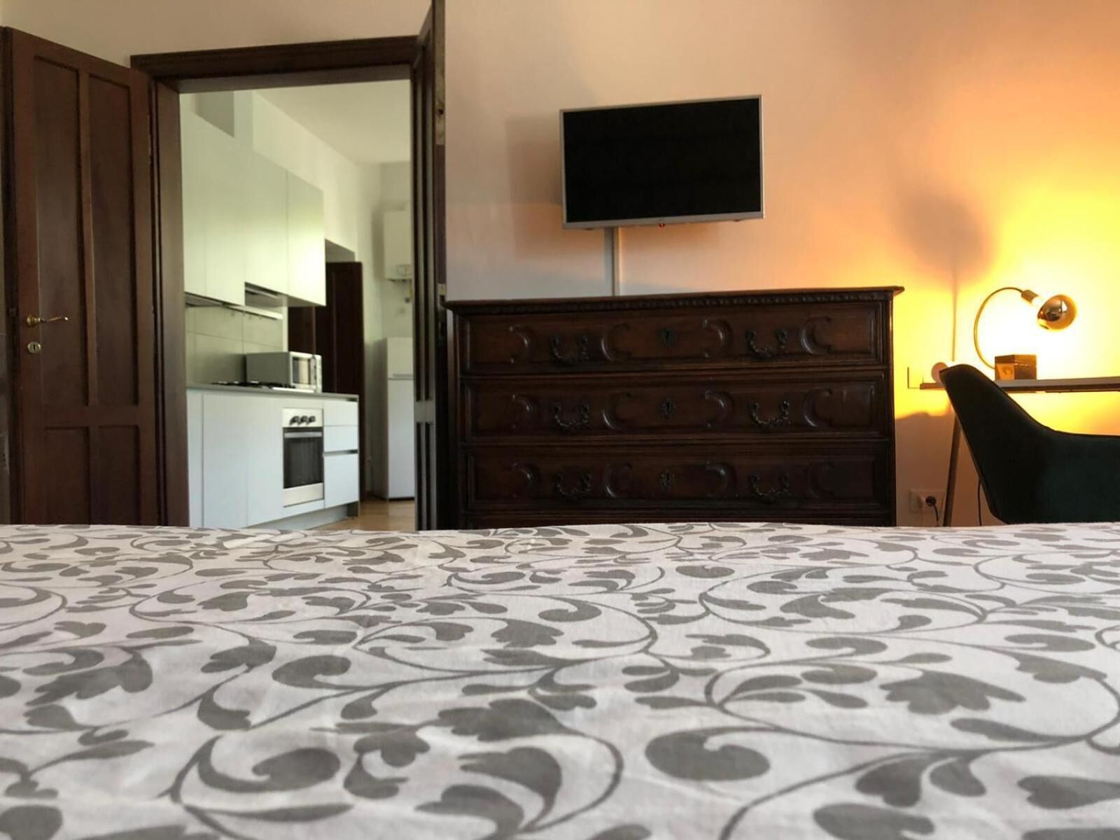 A bedroom with a patterned bedspread, a wooden dresser, a television mounted on the wall, and a desk with a lamp.