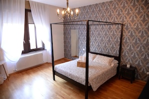 Un lit à baldaquin dans une pièce dont les murs sont recouverts d’un papier peint à motifs, avec un radiateur et une petite table de chevet.