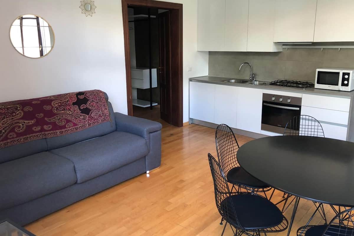 A compact living space with a sofa, a dining table with chairs, a kitchen area including a microwave and stove, and a mirror on the wall.