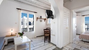 A room with a white table, a chair, a lamp, a small plant, and a bed with a striped bedspread. - Brachera Apartments (Paros)