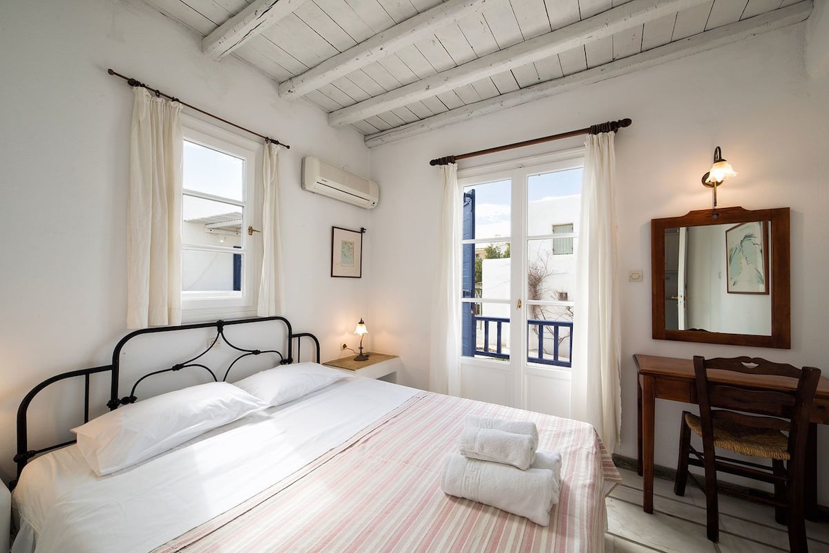 A bedroom with a bed, bedside table, lamp, chair, and a view through a door.