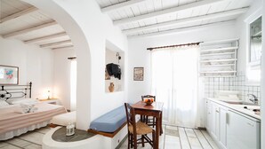 A room with a bed, a small table with a chair, a bench, a window with curtains, and a kitchen area with white cabinets and a sink. - Brachera Apartments (Paros)