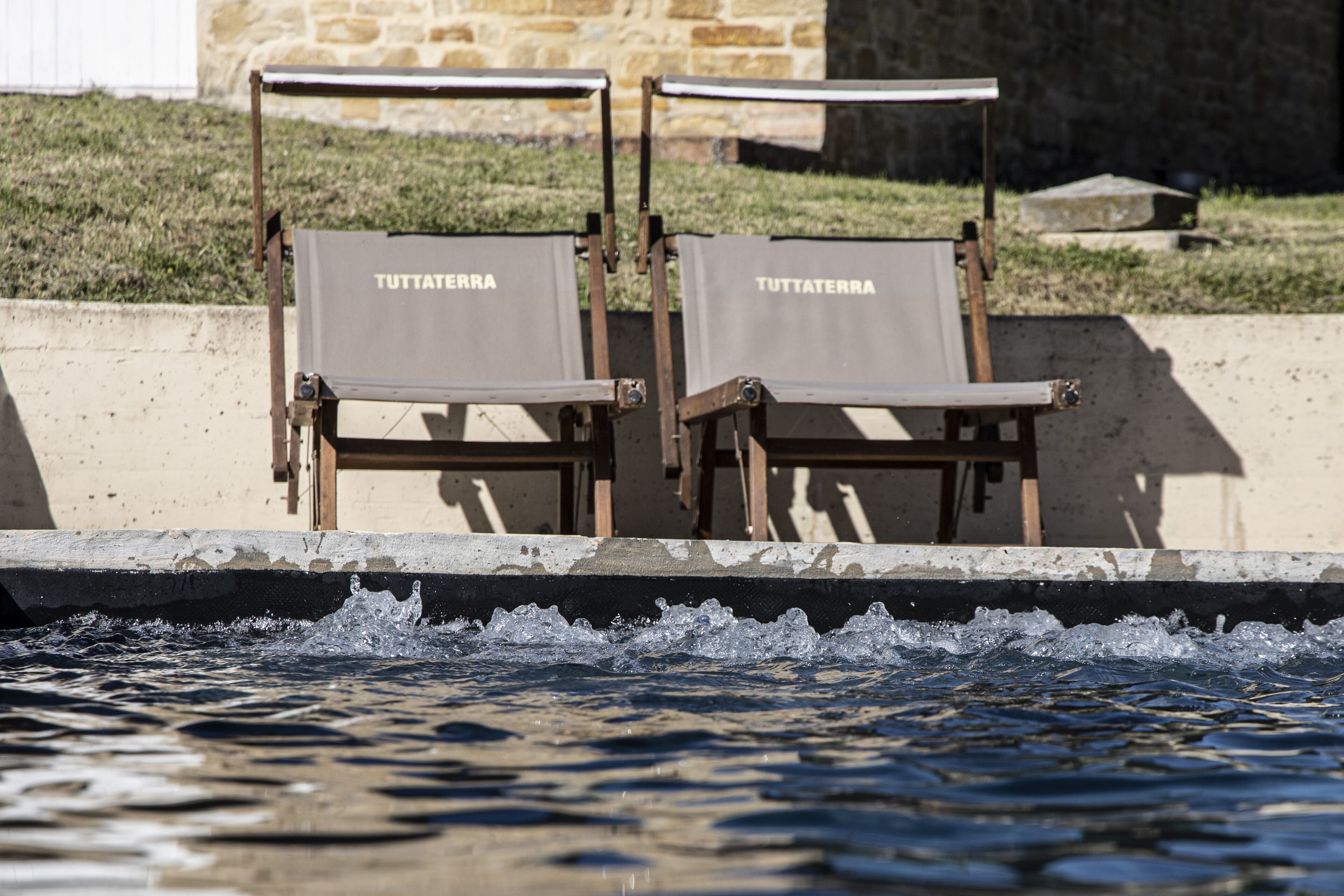 Two TUTTATERRA lounge chairs by a pool with a stone wall and grass in the background.