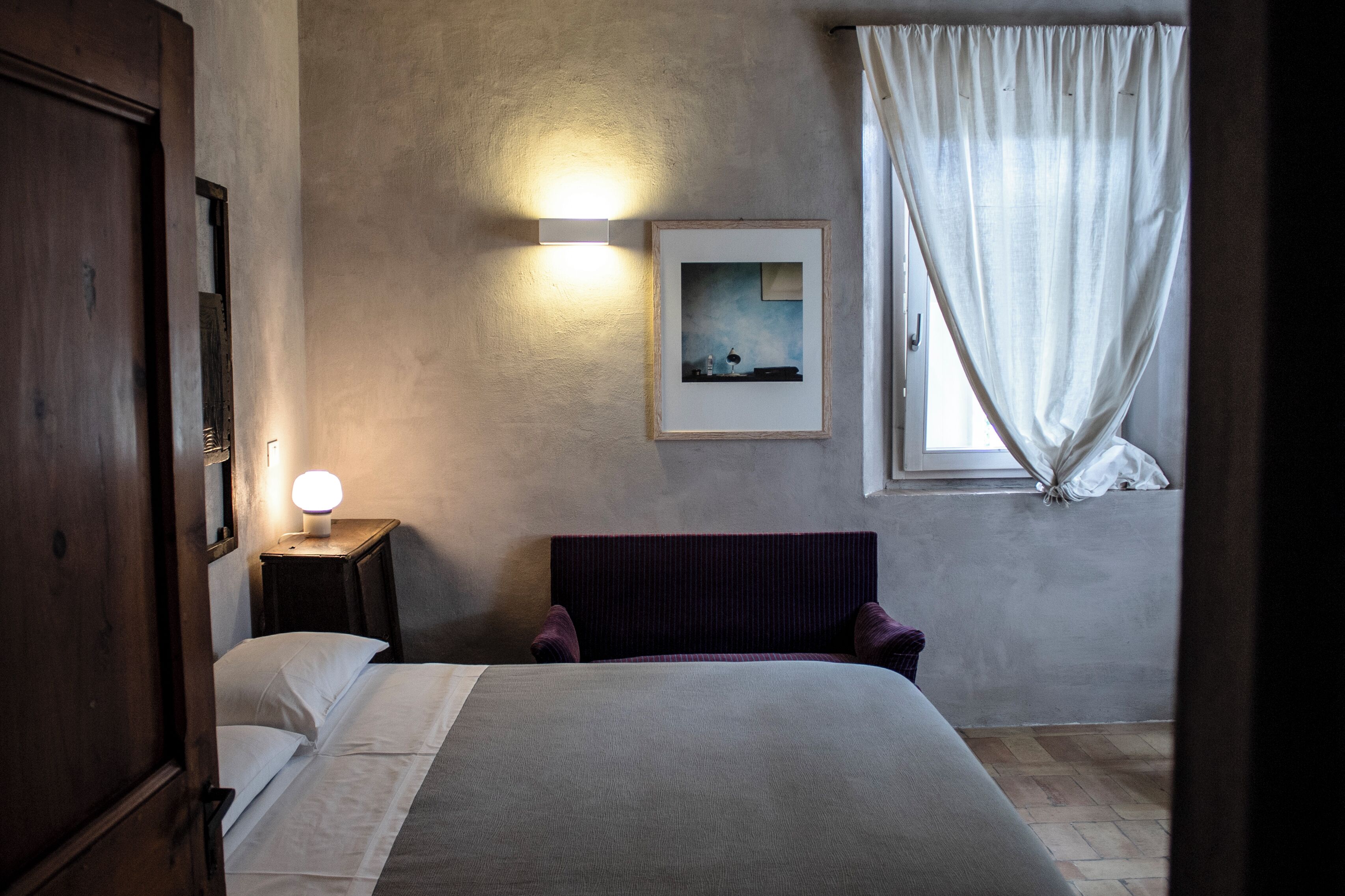 A bedroom with a bed, a bedside table with a lamp, a framed picture on the wall, and a window with curtains.