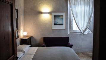 A bedroom with a bed, a bedside table with a lamp, a framed picture on the wall, and a window with curtains.