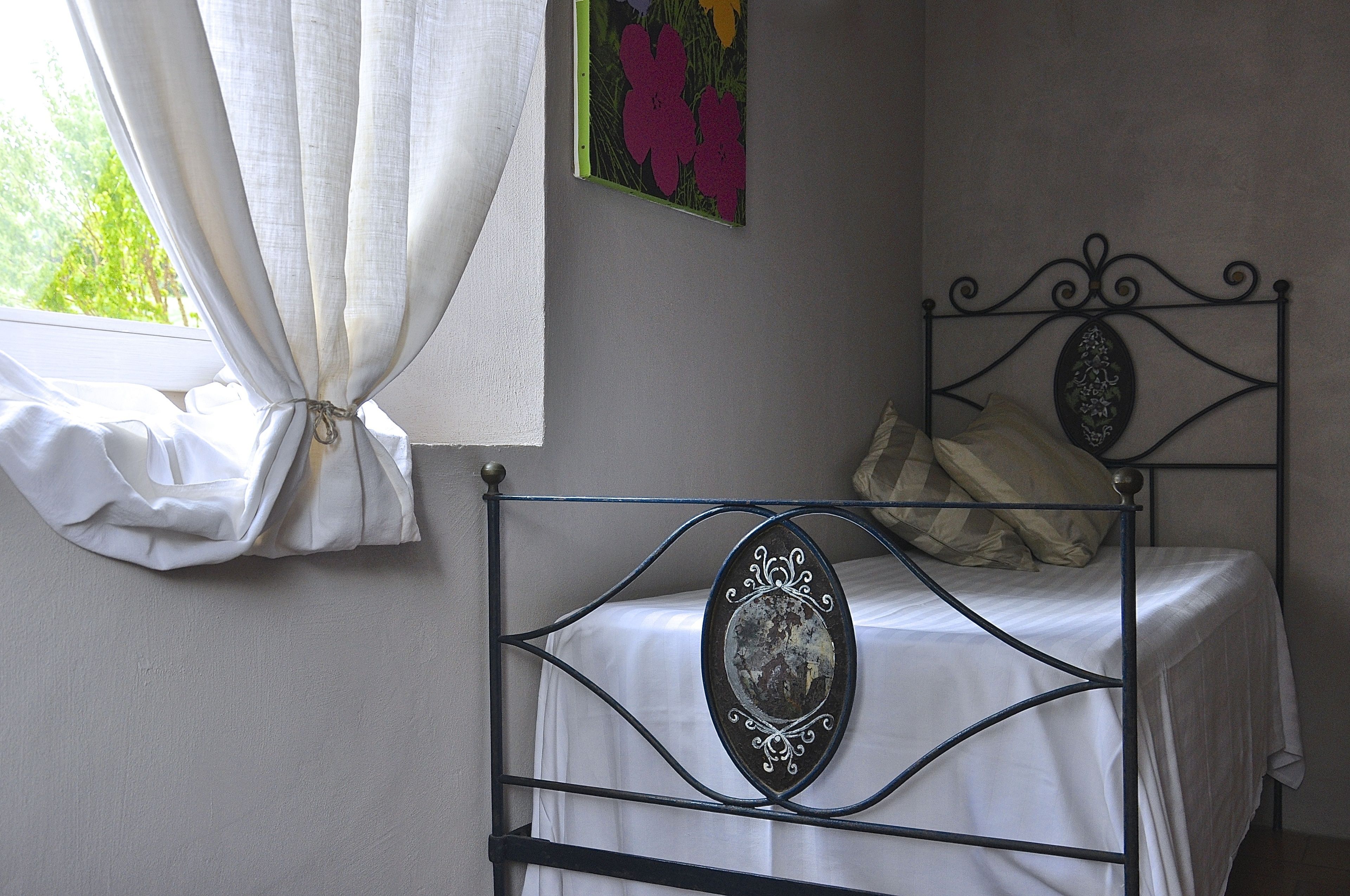 A single bed with a metal frame, white bedding, and a decorative mirror. A window with white curtains is visible, and a colorful painting hangs on the wall.