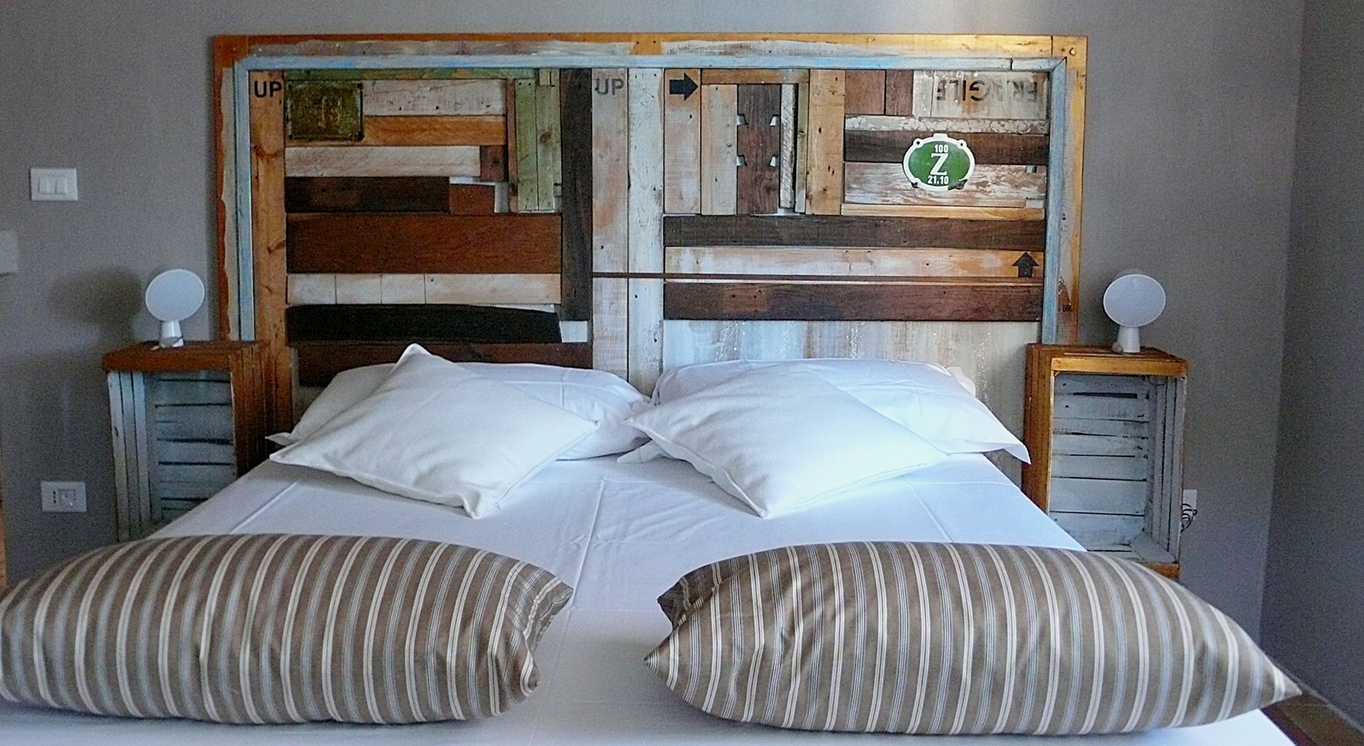 Una cama con sábanas blancas y almohadas a rayas, un cabecero de madera con un reloj, y dos mesitas de noche con lámparas.