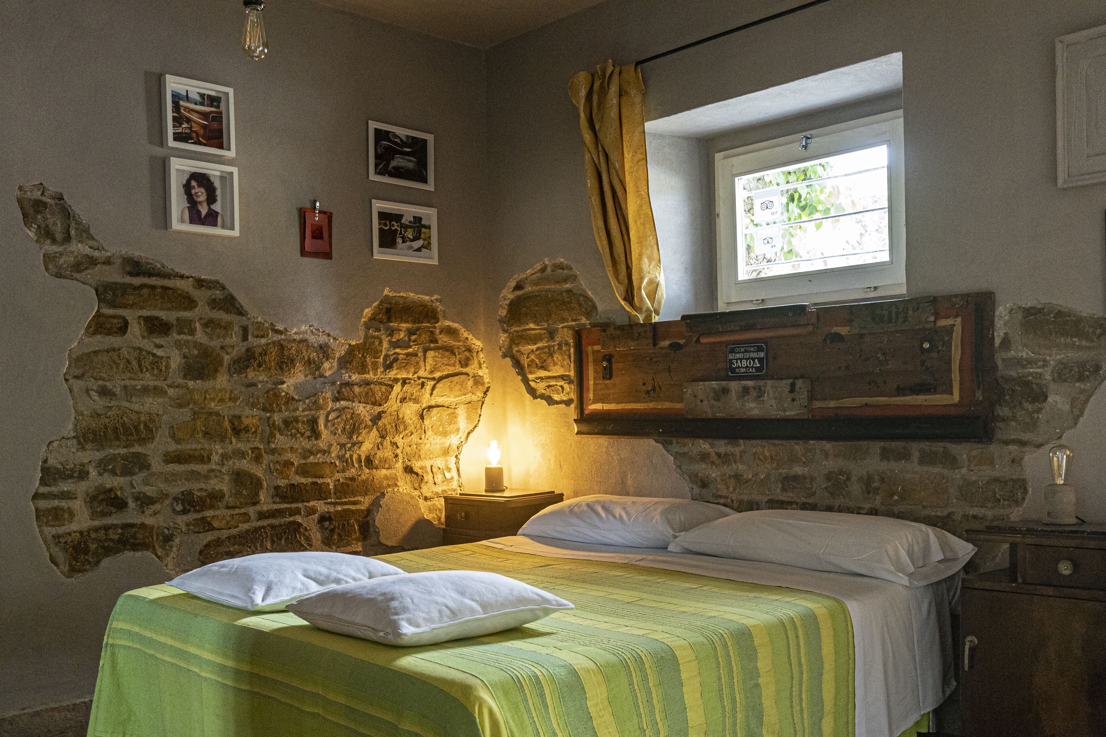Una habitación con una cama, una mesita de noche con una vela, una ventana con cortina y paredes de piedra.