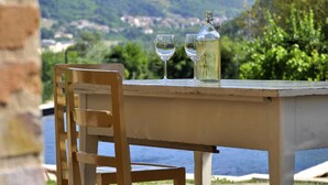 Une table avec deux verres à vin et une bouteille, placée sur un fond pittoresque composé de collines et d’eau.