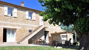 Un bâtiment en pierre avec un escalier menant à un deuxième étage, un espace extérieur aménagé avec des meubles en osier, et un arbre qui offre de l’ombre.