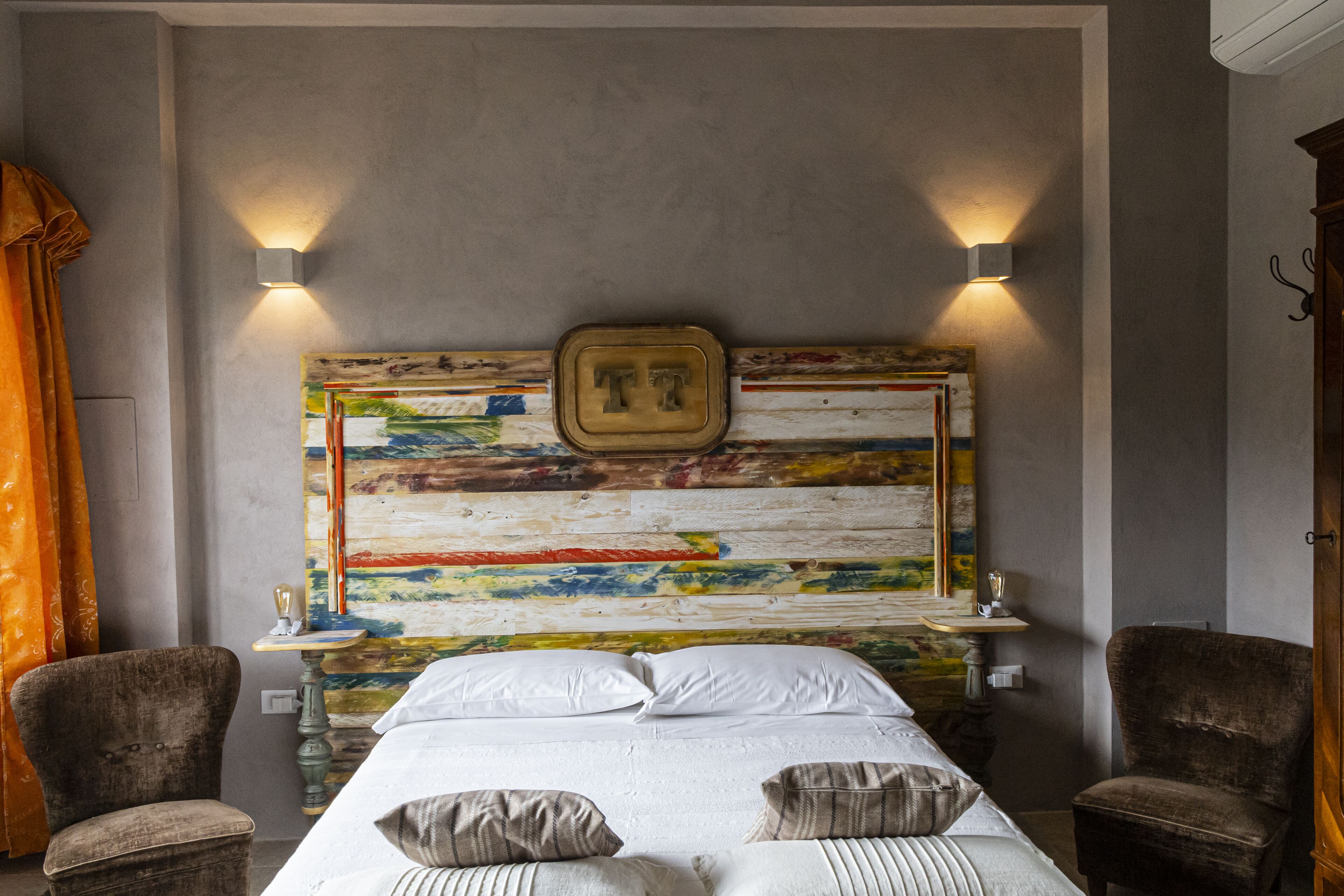 A bed with white linens, two pillows, and a headboard with a colorful abstract design. There are two wall-mounted lights on either side of the headboard.