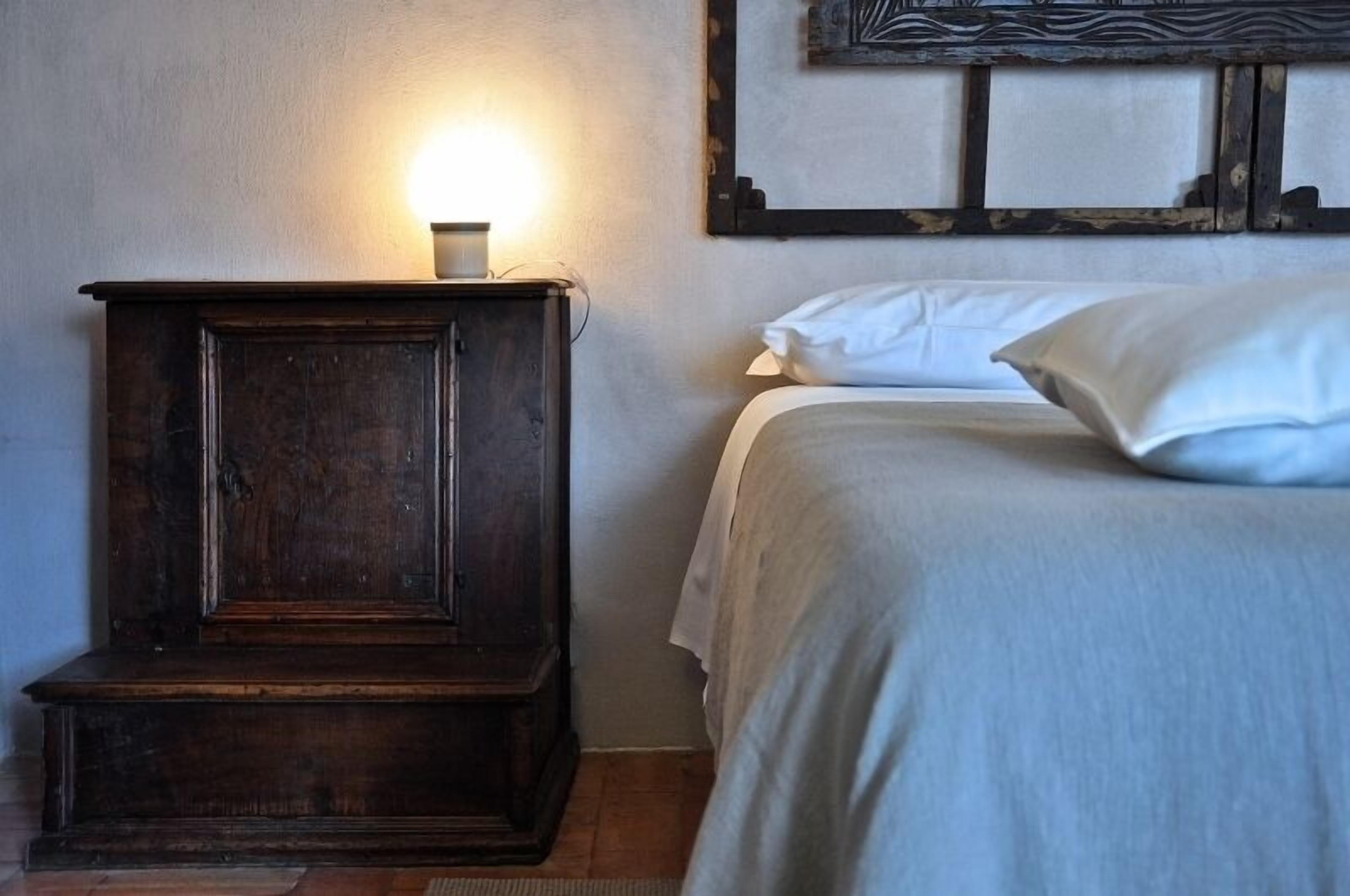 A bedroom with a wooden nightstand, a bedside lamp, a bed with white bedding, and a wall-mounted artwork.