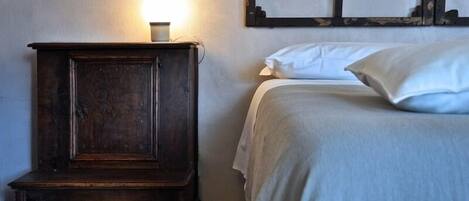 Une chambre avec une table de chevet en bois, une lampe de chevet, un lit avec une literie blanche et une œuvre d’art accrochée au mur.