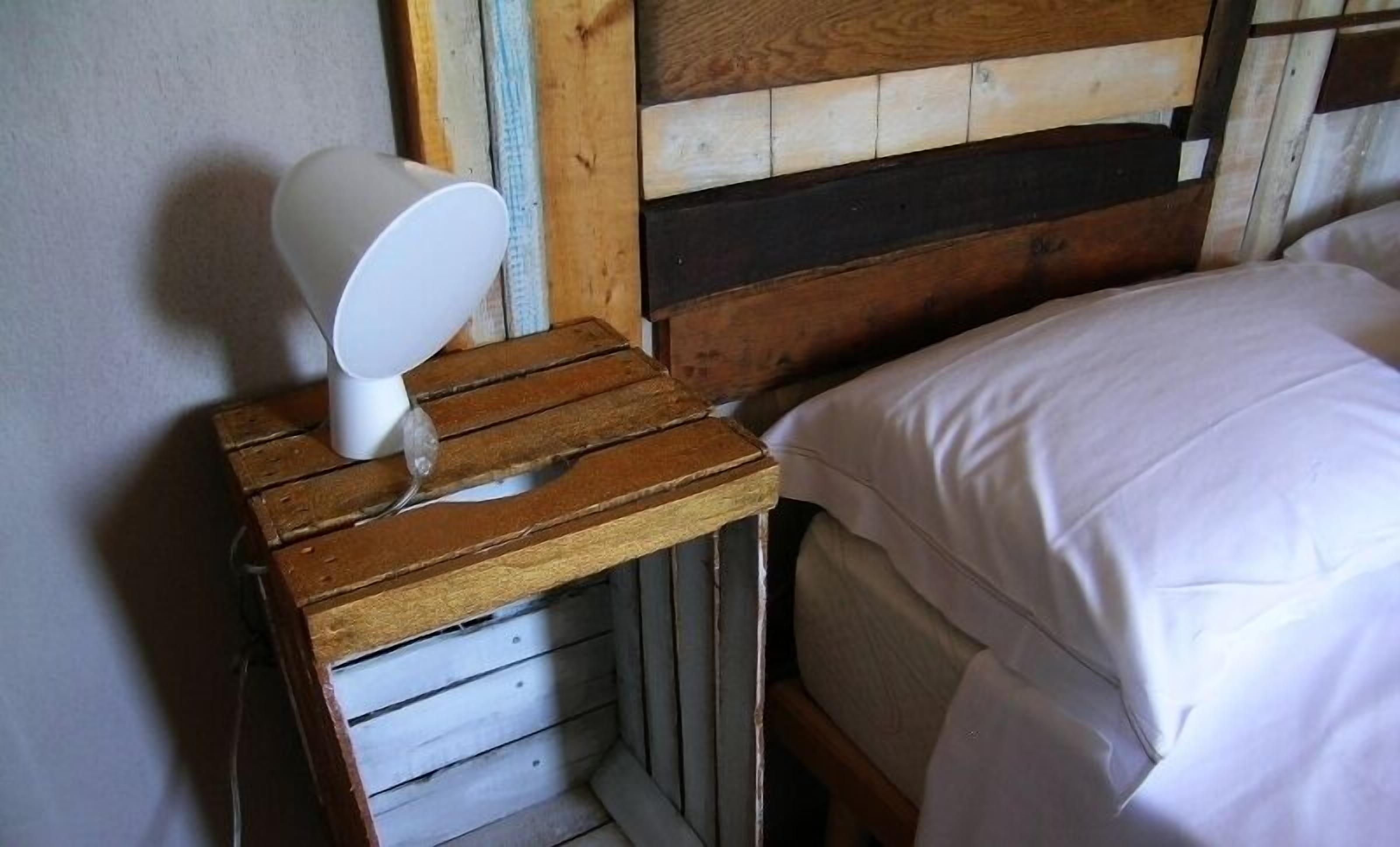 A wooden bedside table with a white lamp, a wooden headboard, and a white bedspread.
