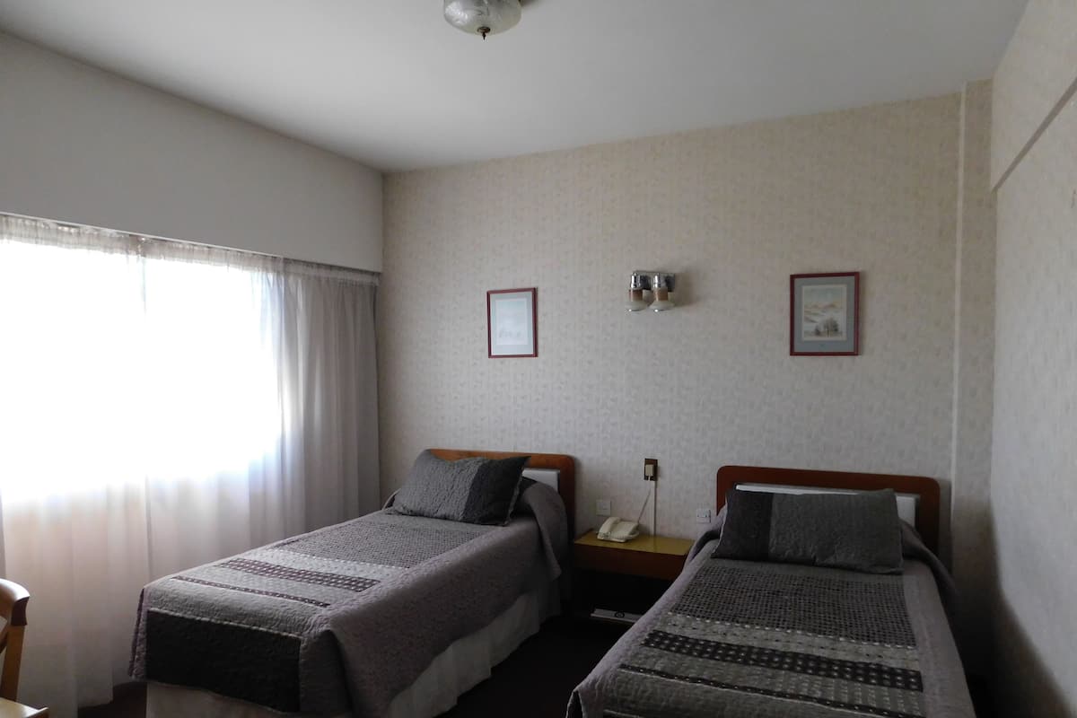 A hotel room with two single beds, a bedside table with a lamp, a window with curtains, and a wall-mounted picture.