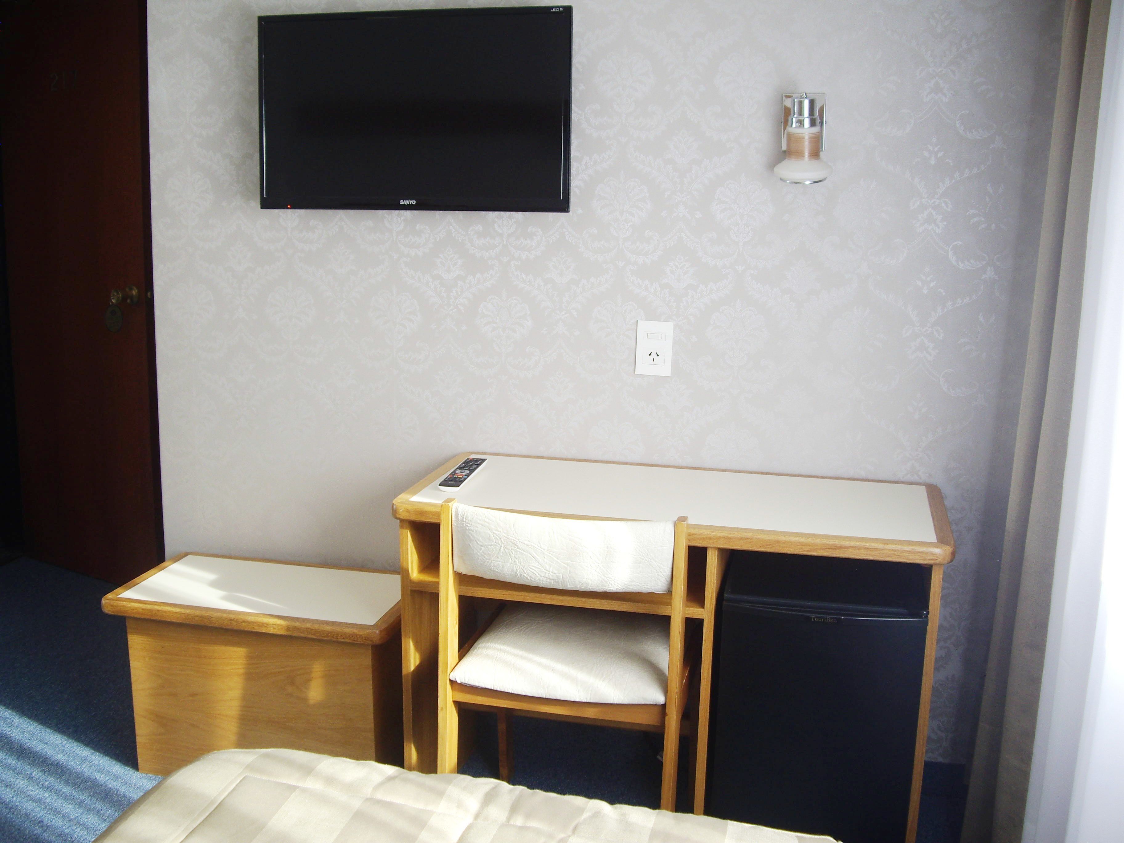 Habitación compacta con cama, escritorio y silla, televisor montado en la pared y una mesita de noche pequeña.